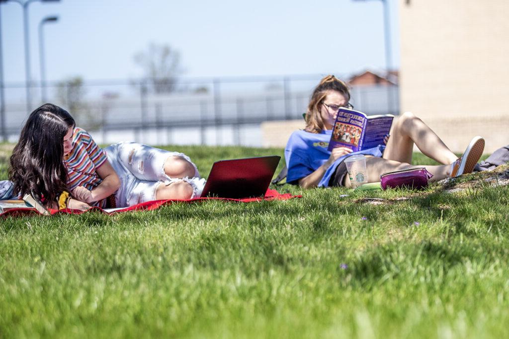 Two people lying on the grass; one is reading a book and the other is using a laptop.